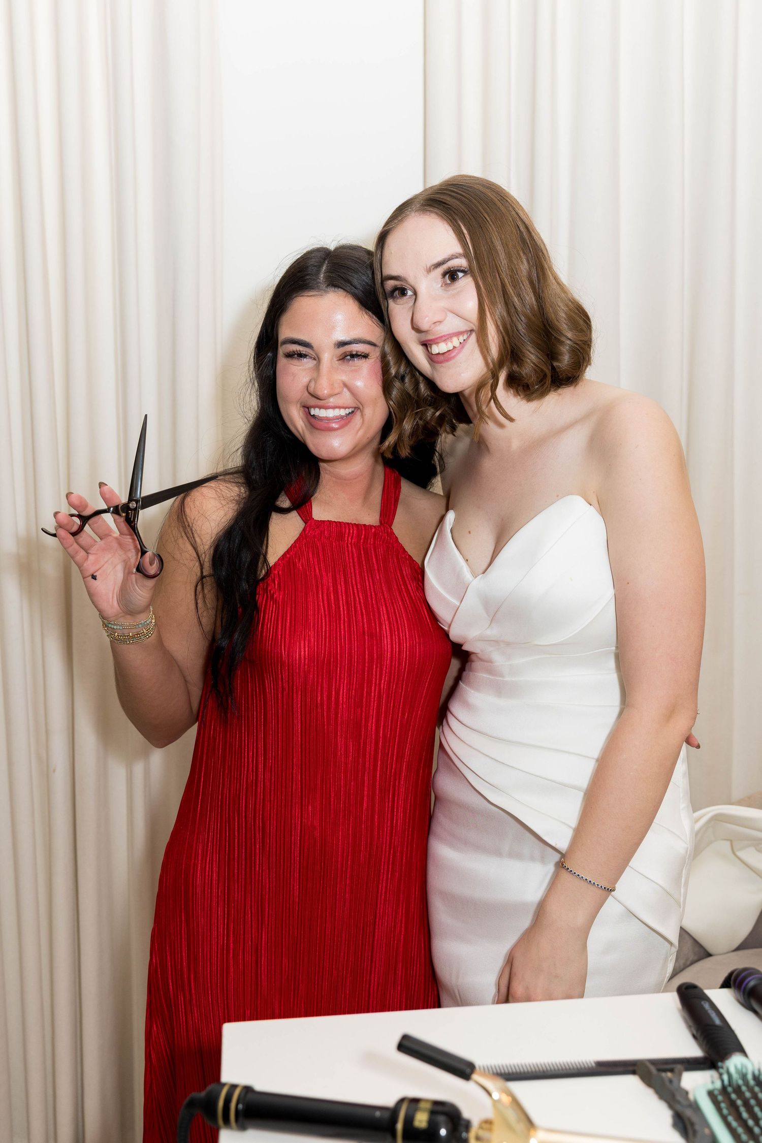 Two women smile together, one in red dress holding scissors, other in white gown, with hair tools on table nearby.