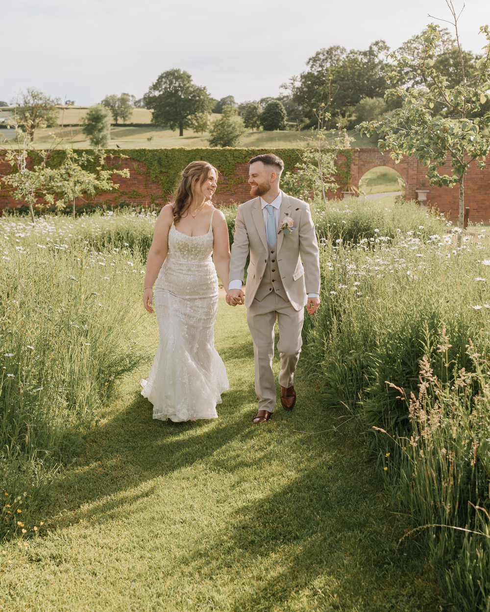 A bride and groom walk hand in hand through a lush garden path on their wedding day in the English countryside.