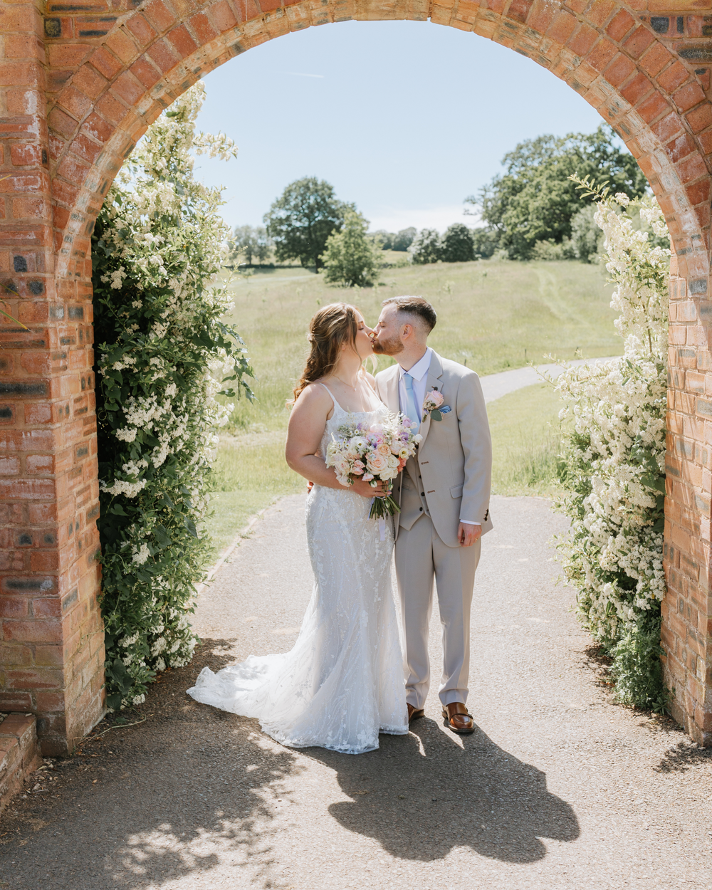 Bride and groom kissing under a brick arch adorned with white flowers at an outdoor wedding venue.