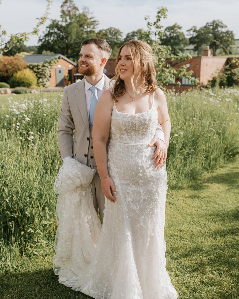 A bride in a lace gown and groom in a tan suit stand smiling in a lush green garden on their wedding day.