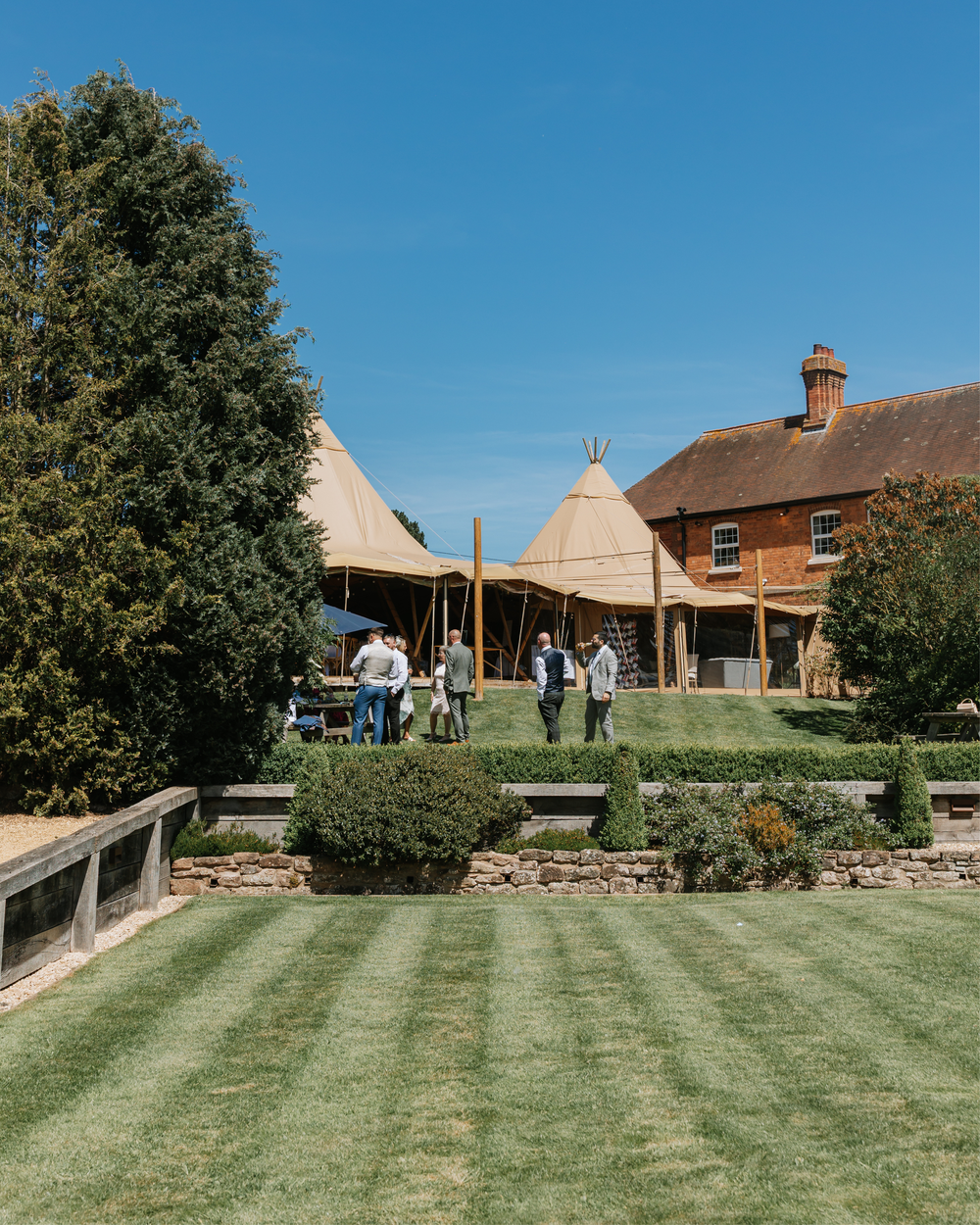 Elegant countryside wedding venue with tipi tents, manicured lawn, and brick manor house under clear blue sky.