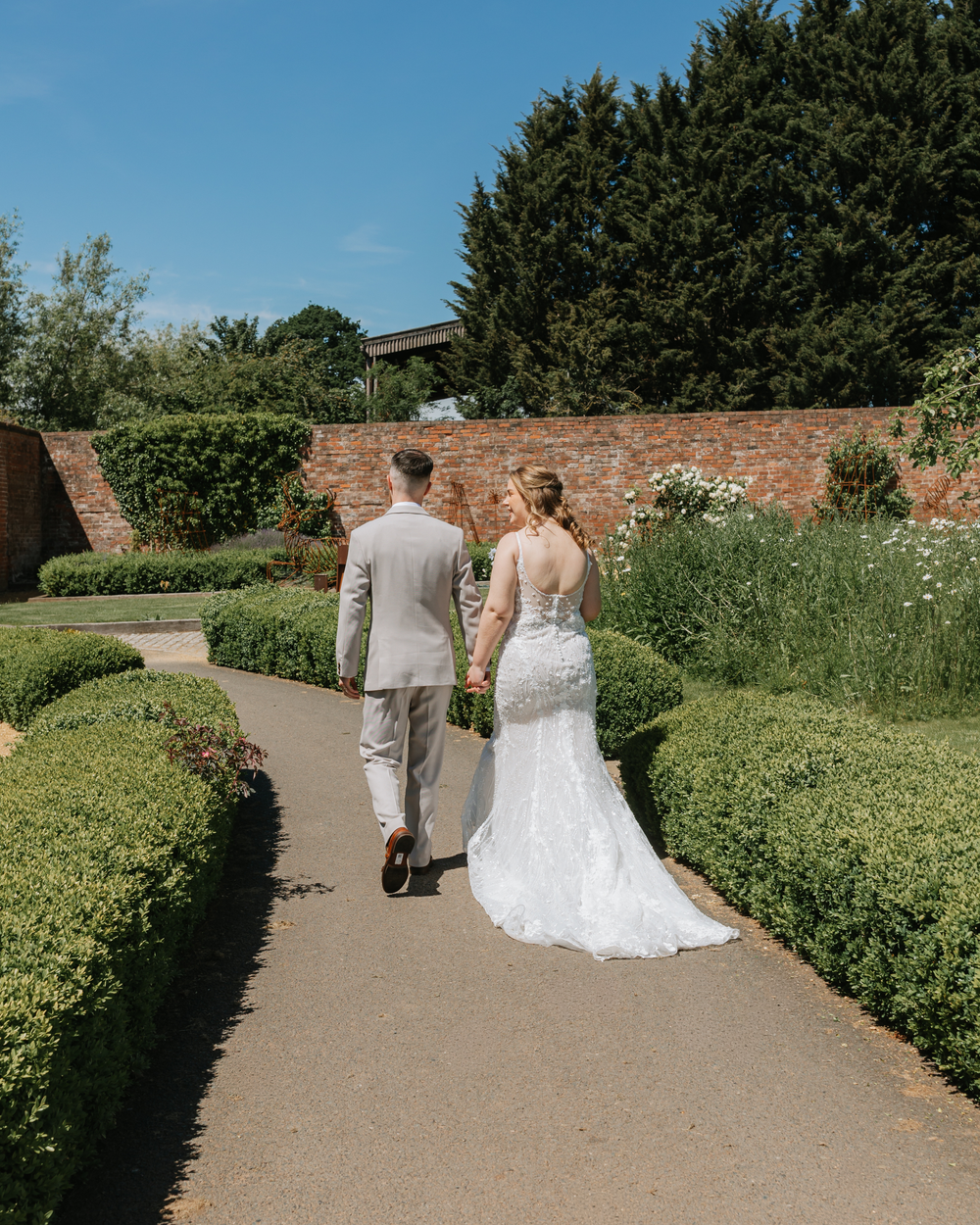 A bride and groom walk hand in hand through a lush garden pathway on their wedding day.