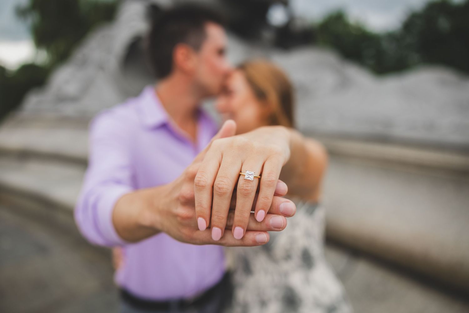 Couple sharing a kiss while showing off an engagement ring with pink nails in a romantic outdoor setting.