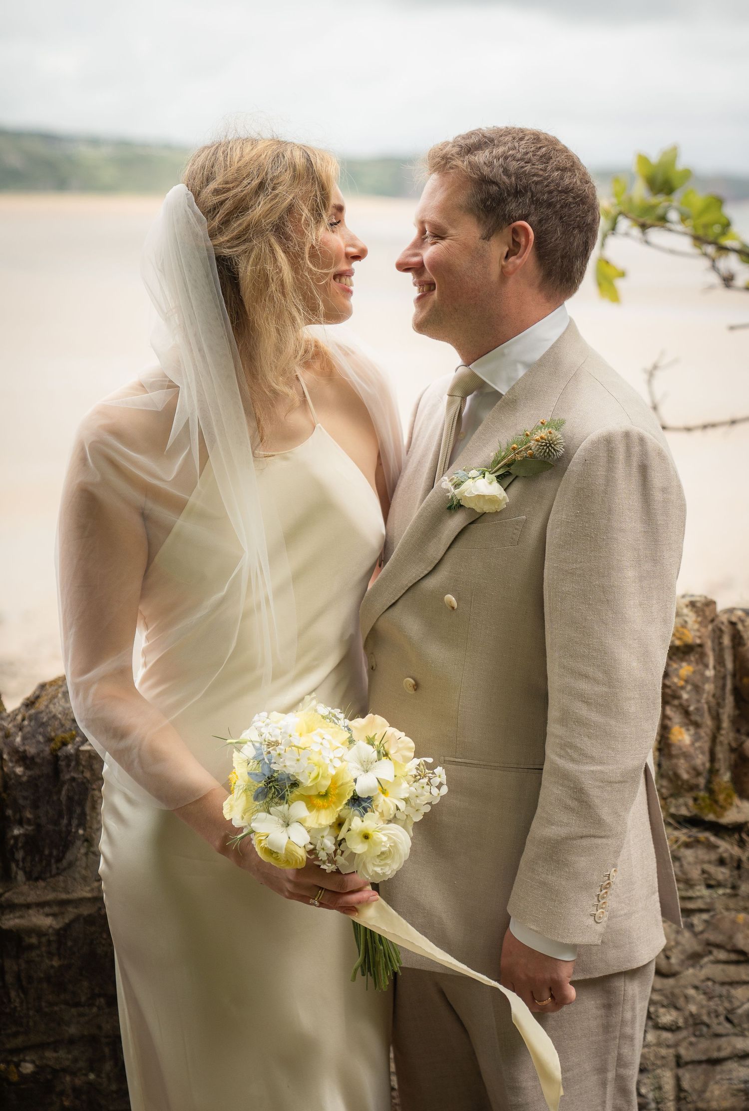 Bride and groom share a romantic gaze outdoors, she holds yellow bouquet, both in elegant cream wedding attire.