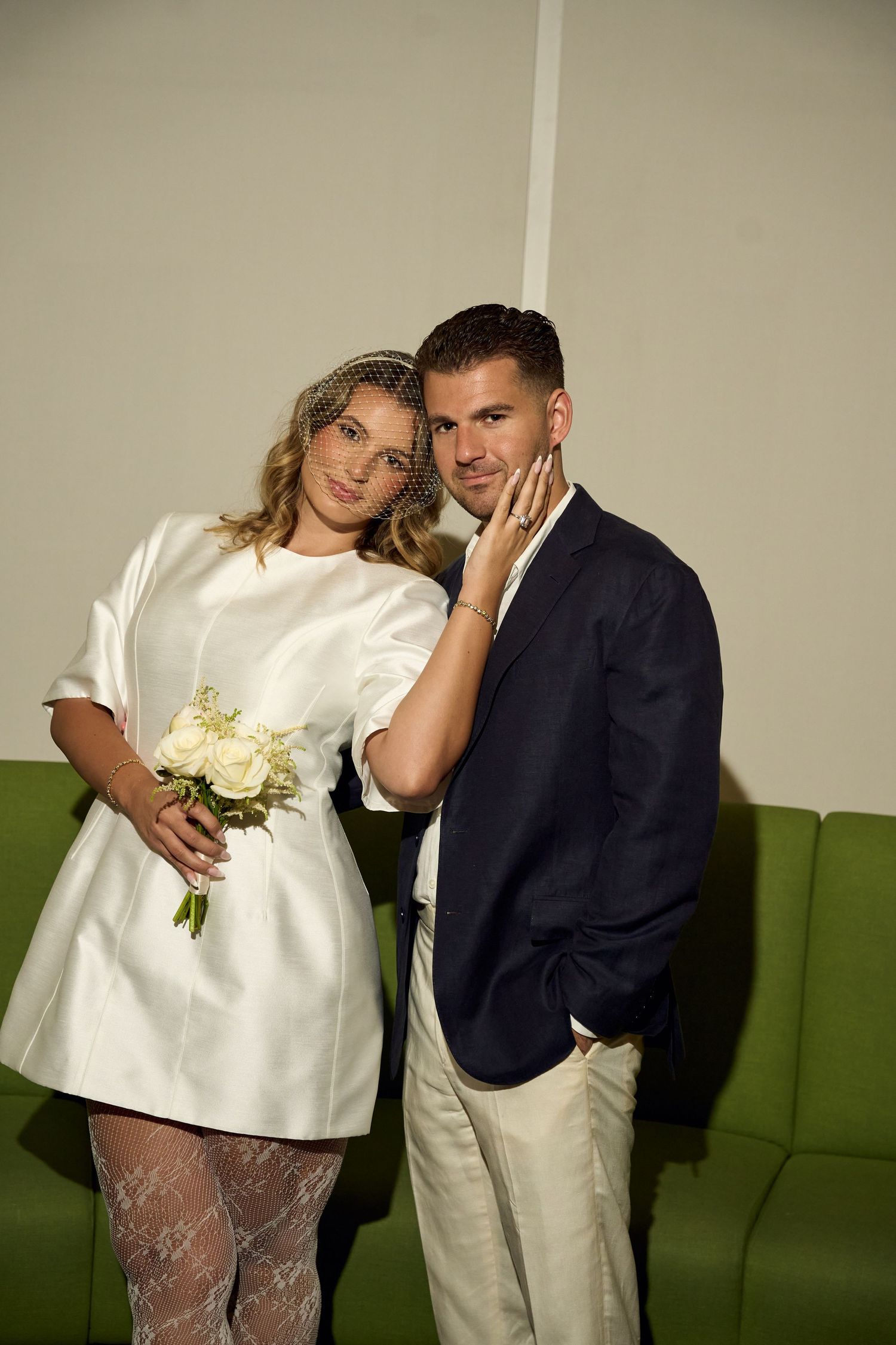 Elegant bride in white mini dress holding bouquet, groom in navy suit, romantic wedding portrait pose.