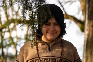 Smiling person wearing a patterned sweater and dark hat with pine branches, standing outdoors among trees.
