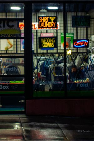 Neon signs for shirt laundry and wedding gowns glow in a storefront window at night, with an open sign visible.