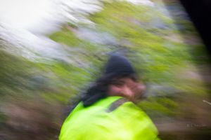 Blurred motion photo of a person wearing a neon yellow jacket and black beanie outdoors near green trees.