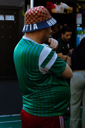 Man wearing Croatia bucket hat and green Mexico soccer jersey standing outdoors at a sports event.