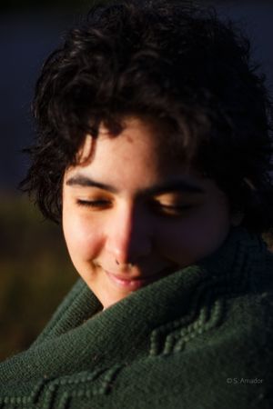 A young person with curly dark hair smiling softly, wrapped in a green knit sweater in warm sunlight.