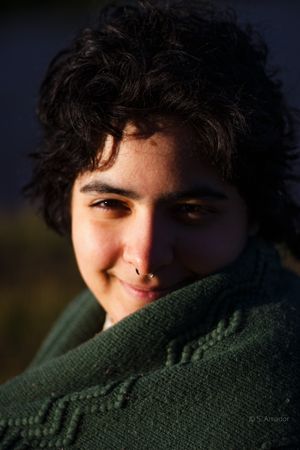 Young woman with curly dark hair and nose ring smiling, wrapped in green sweater.
