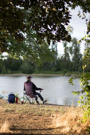 Person sitting in a folding chair fishing by a calm lake, surrounded by lush green trees on a sunny day.
