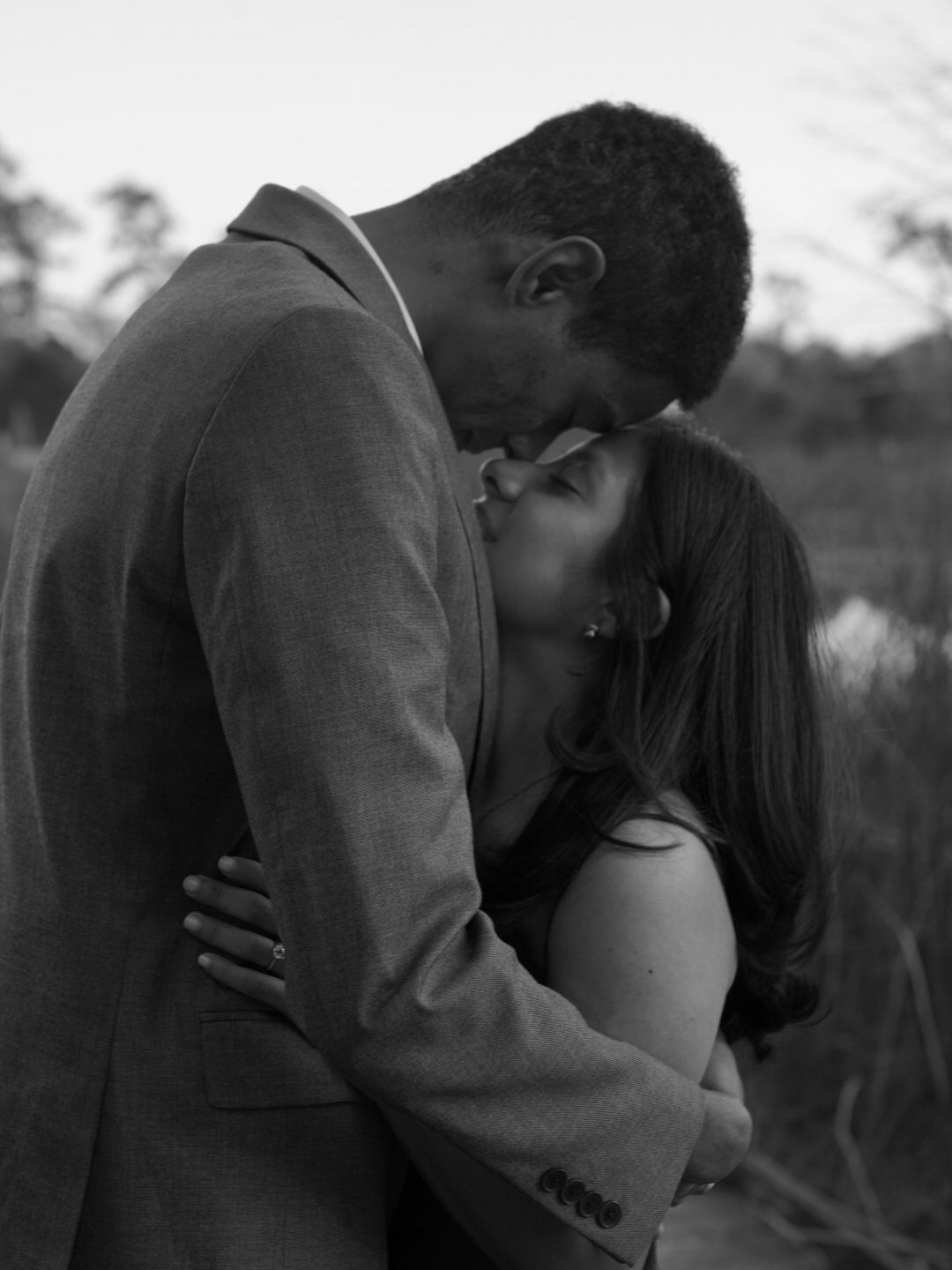 Black and white photo of a couple sharing a romantic kiss outdoors, man in suit jacket embracing woman.