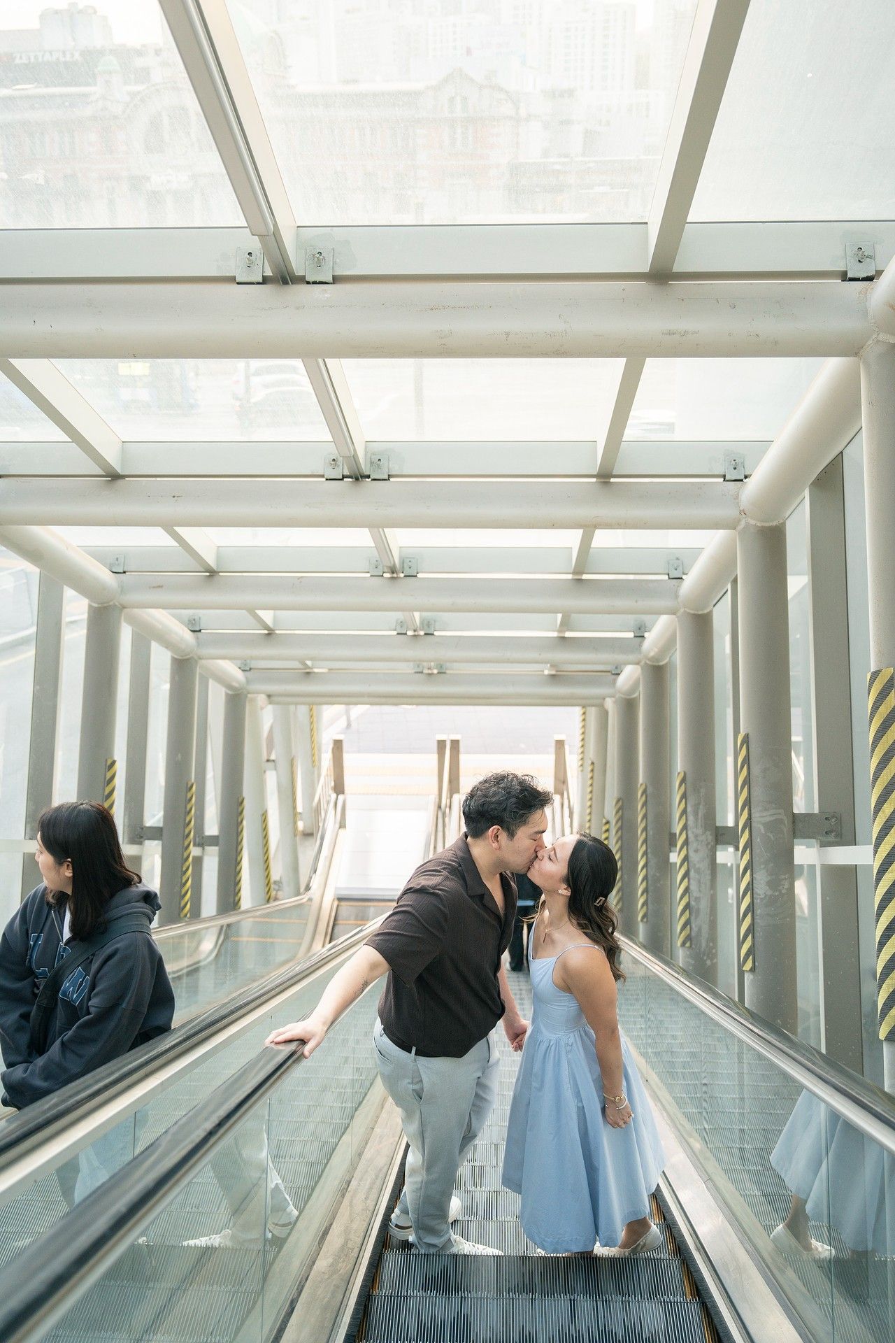 Couple sharing a romantic kiss on an escalator inside a modern glass-roofed walkway, with another person visible nearby.