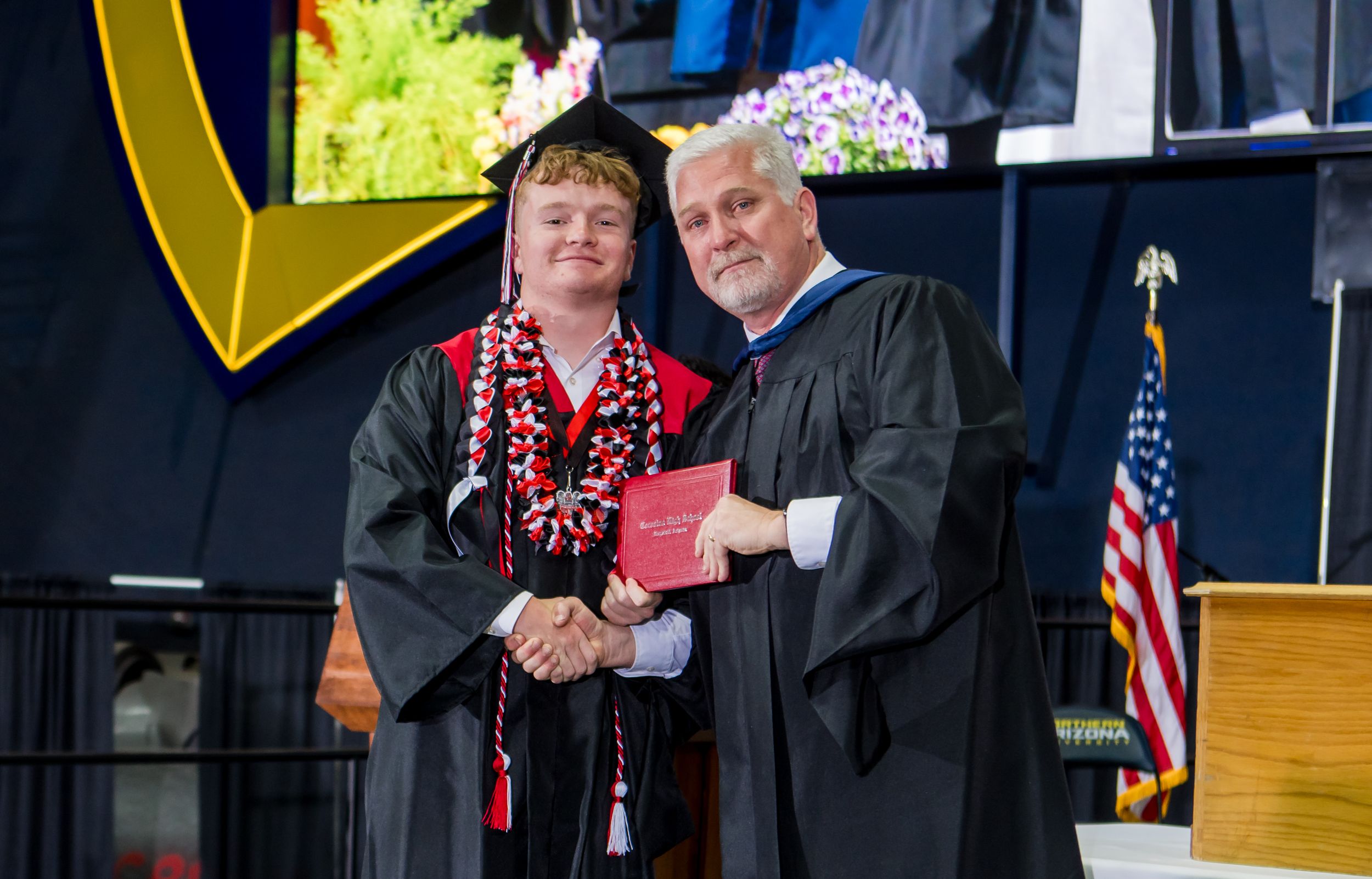 A graduate student in cap and gown receives their diploma from faculty member during commencement ceremony on stage.