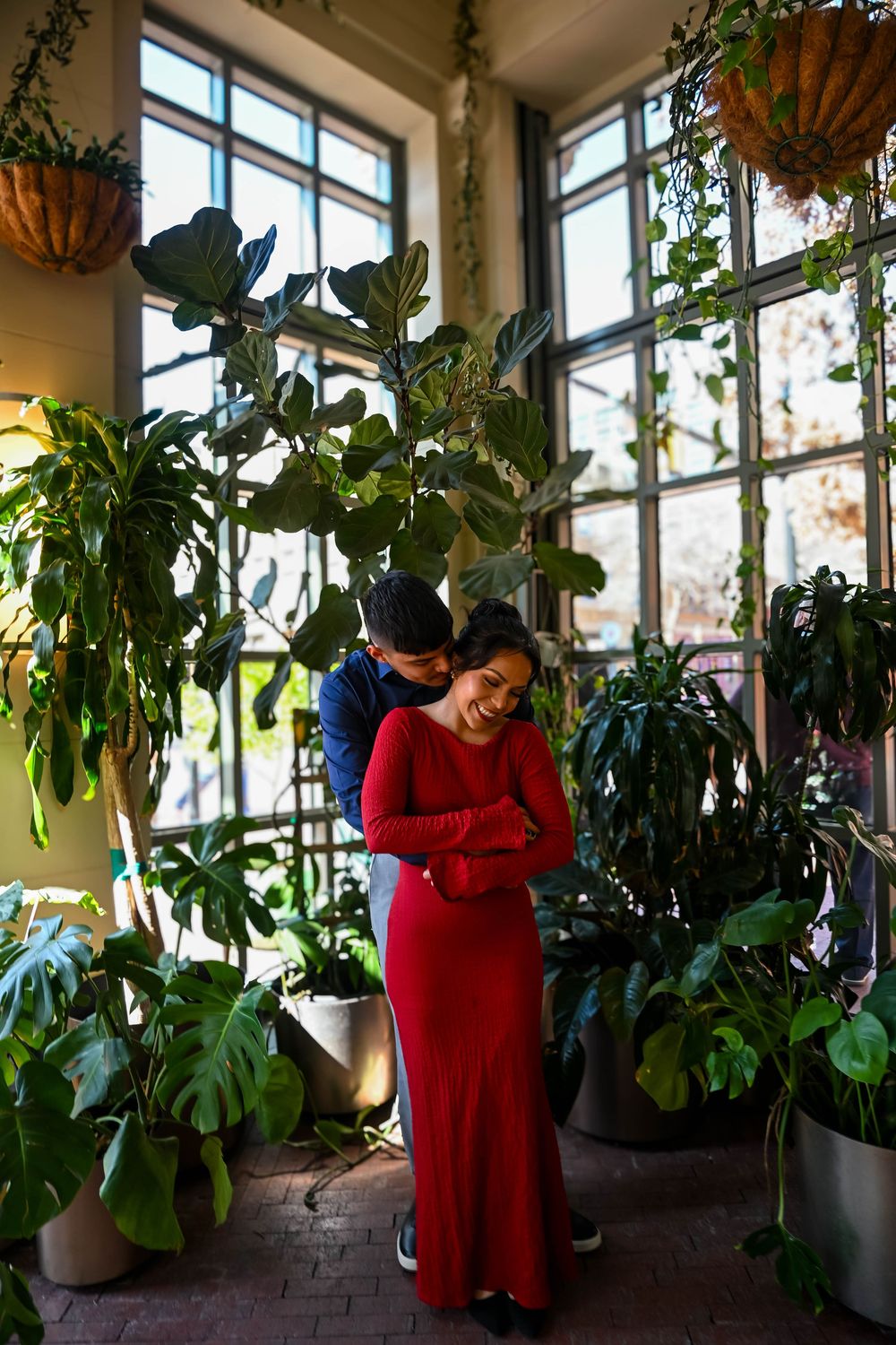 Couple embracing among lush indoor tropical plants near large windows in a greenhouse setting, woman in red dress.