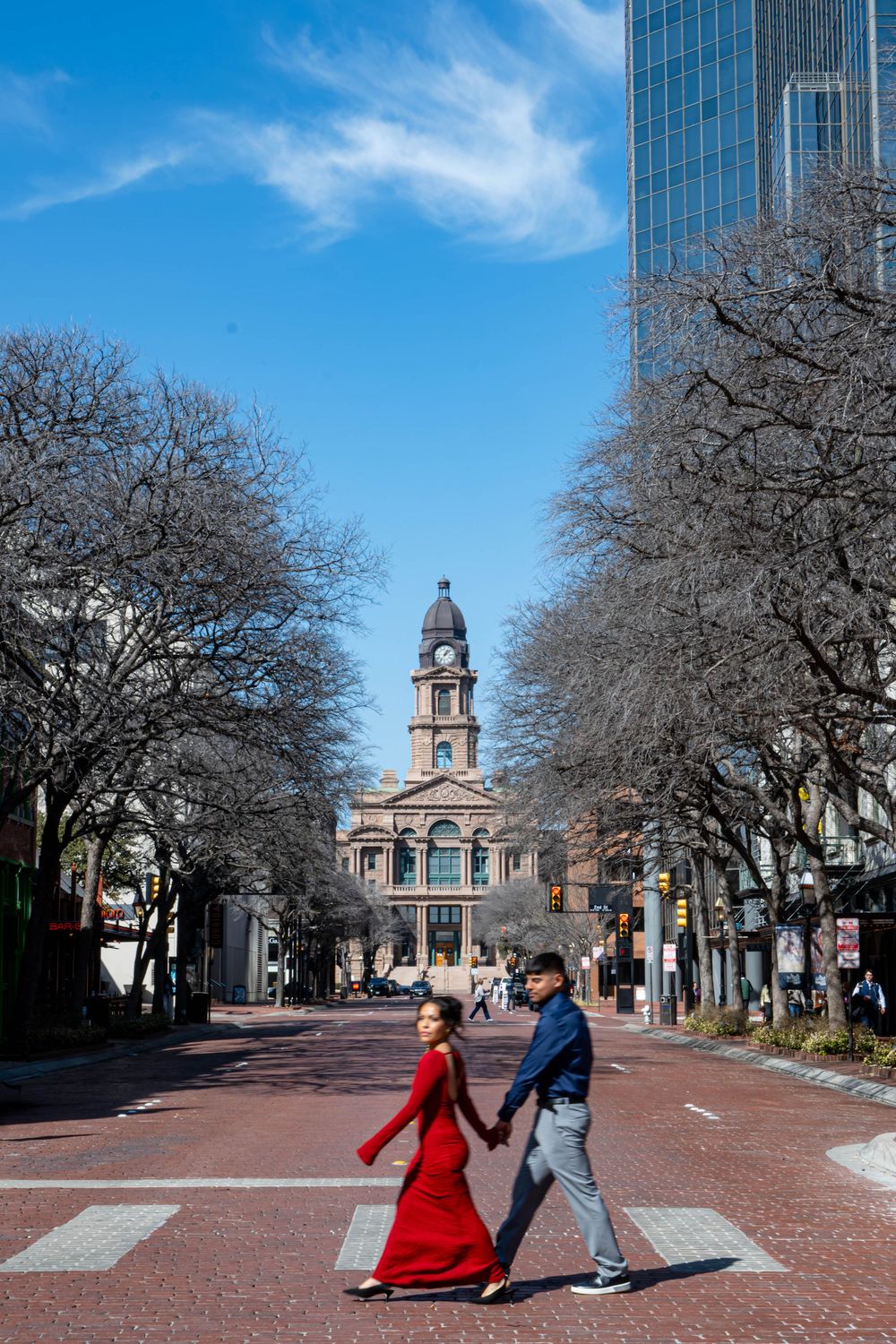 Couple holding hands crossing a brick pedestrian street with a historic clock tower and blue sky in Fort Worth, TX.