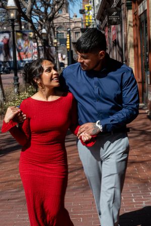 Smiling couple walking together on a sunny brick sidewalk, woman in red dress and man in blue shirt.