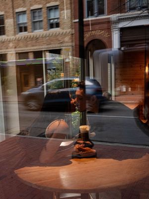 Artistic window reflection shot of a coffee shop interior with a wooden table, drink, and city street outside.