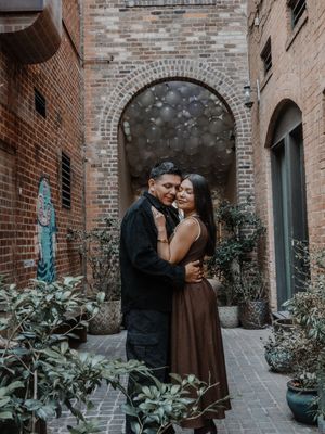 Couple embracing in a charming brick courtyard with an arched doorway and lush green plants surrounding them.