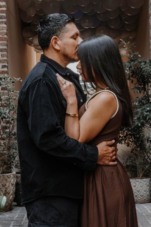 Couple sharing a romantic kiss in an outdoor courtyard with brick walls and potted plants in the background.