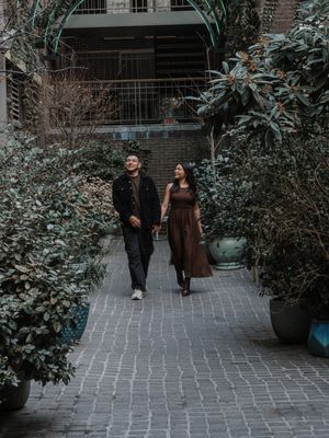 Couple walking hand in hand along a brick pathway lined with lush green shrubs and potted plants outdoors.