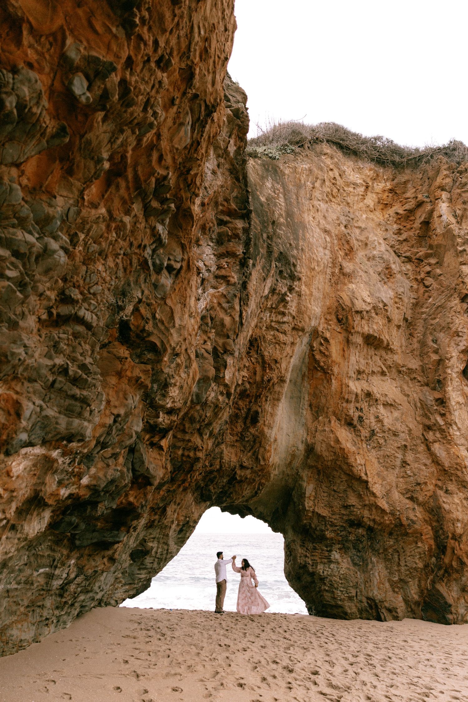 Couple standing under a natural rock arch on a sandy beach with dramatic orange cliffs framing the ocean view.