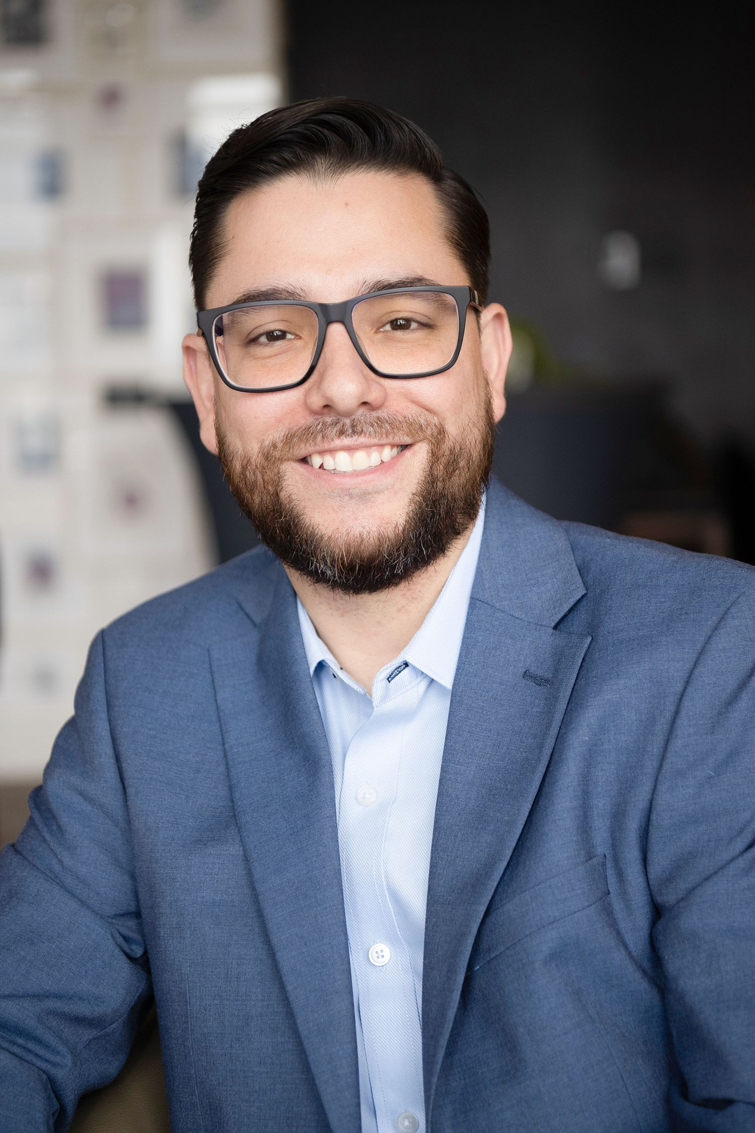 Smiling bearded man with glasses wearing a blue blazer and light blue shirt in a professional office setting.
