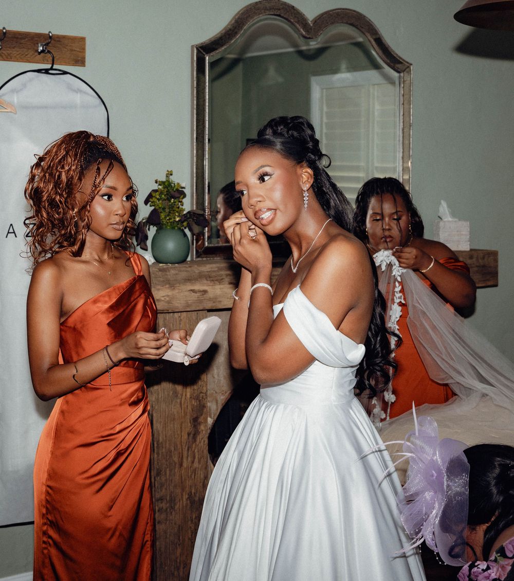 Bridesmaids in burnt orange dresses help bride in white gown prepare for wedding ceremony.