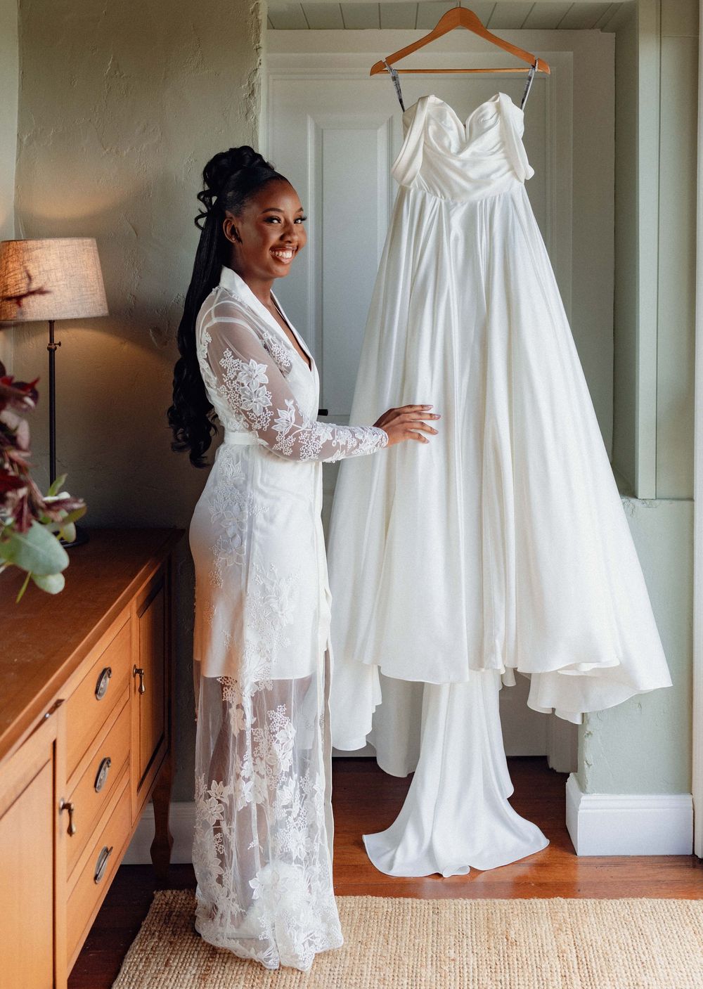 Beautiful white wedding gowns displayed in a room with a wooden dresser and lamp, showcasing bridal fashion.