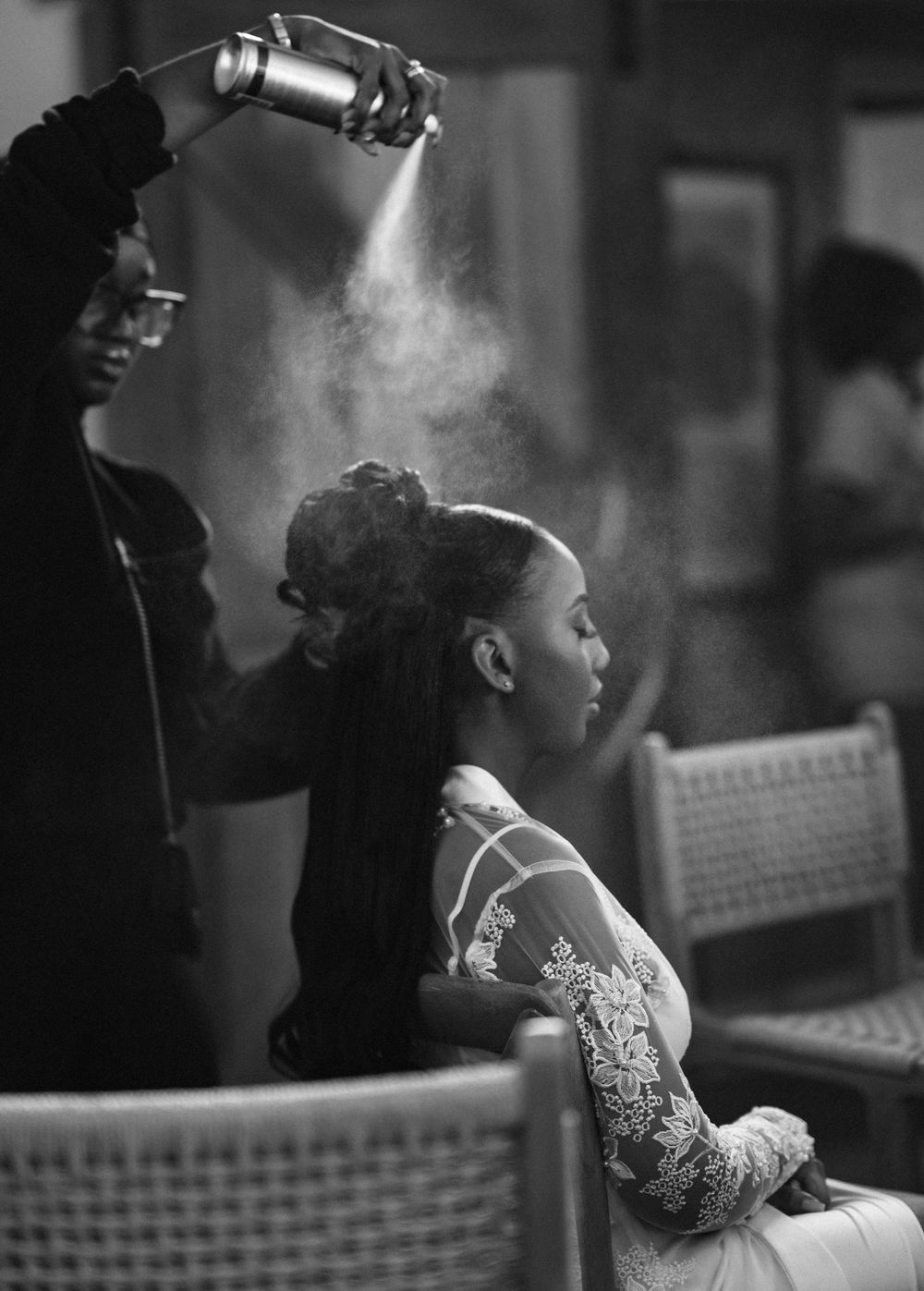 A stylist applies hairspray in a dramatic black and white shot showing mist illuminated against a dark background.