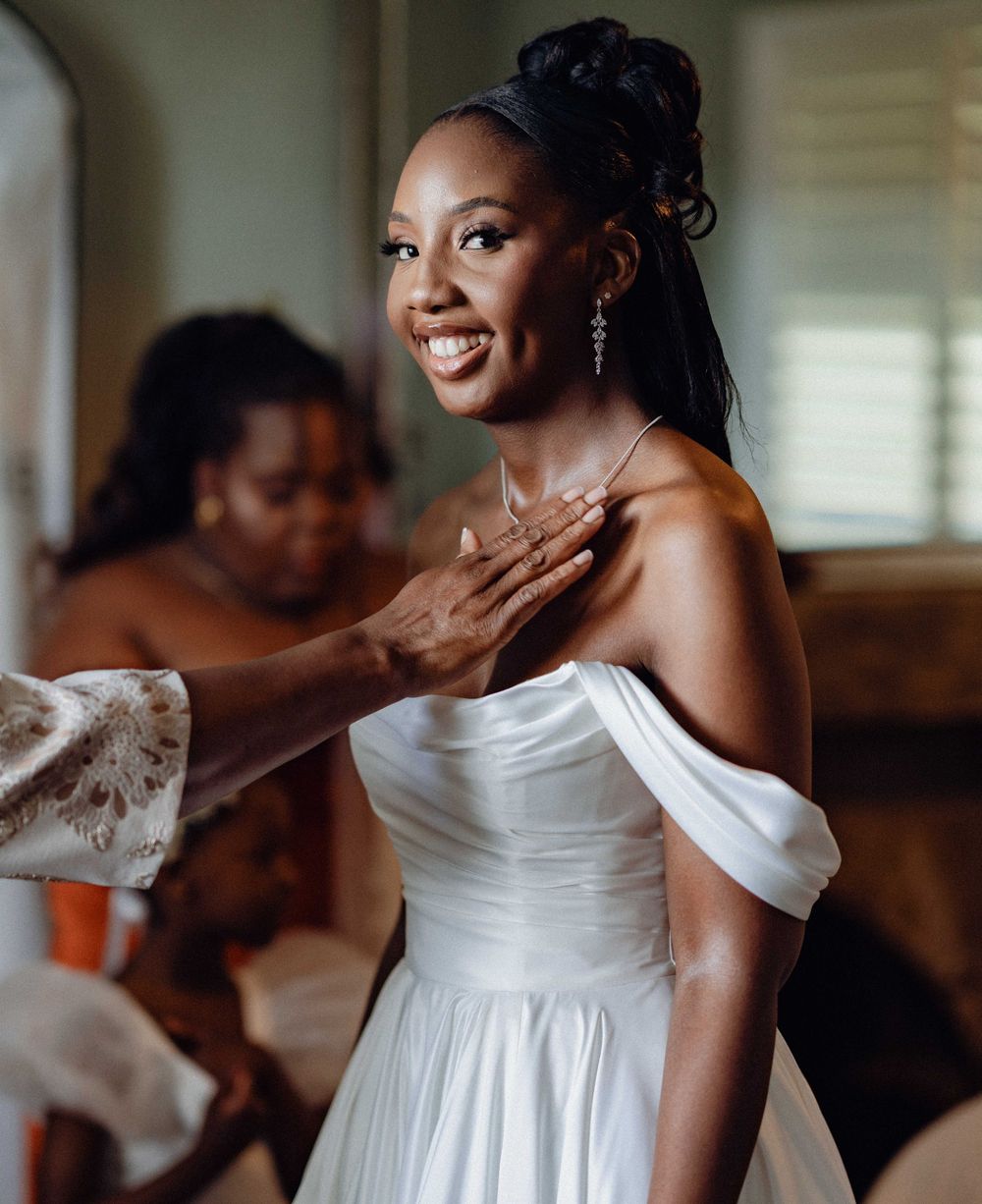 A person in a white off-shoulder wedding dress smiles joyfully while getting ready in a softly lit room.