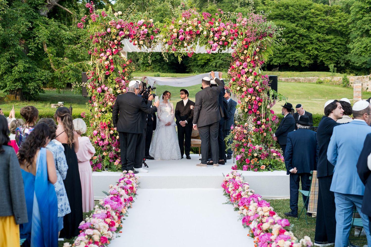 Vue large d’une Houppa décorée de fleurs, mariés et famille réunis pendant un mariage juif en plein air.