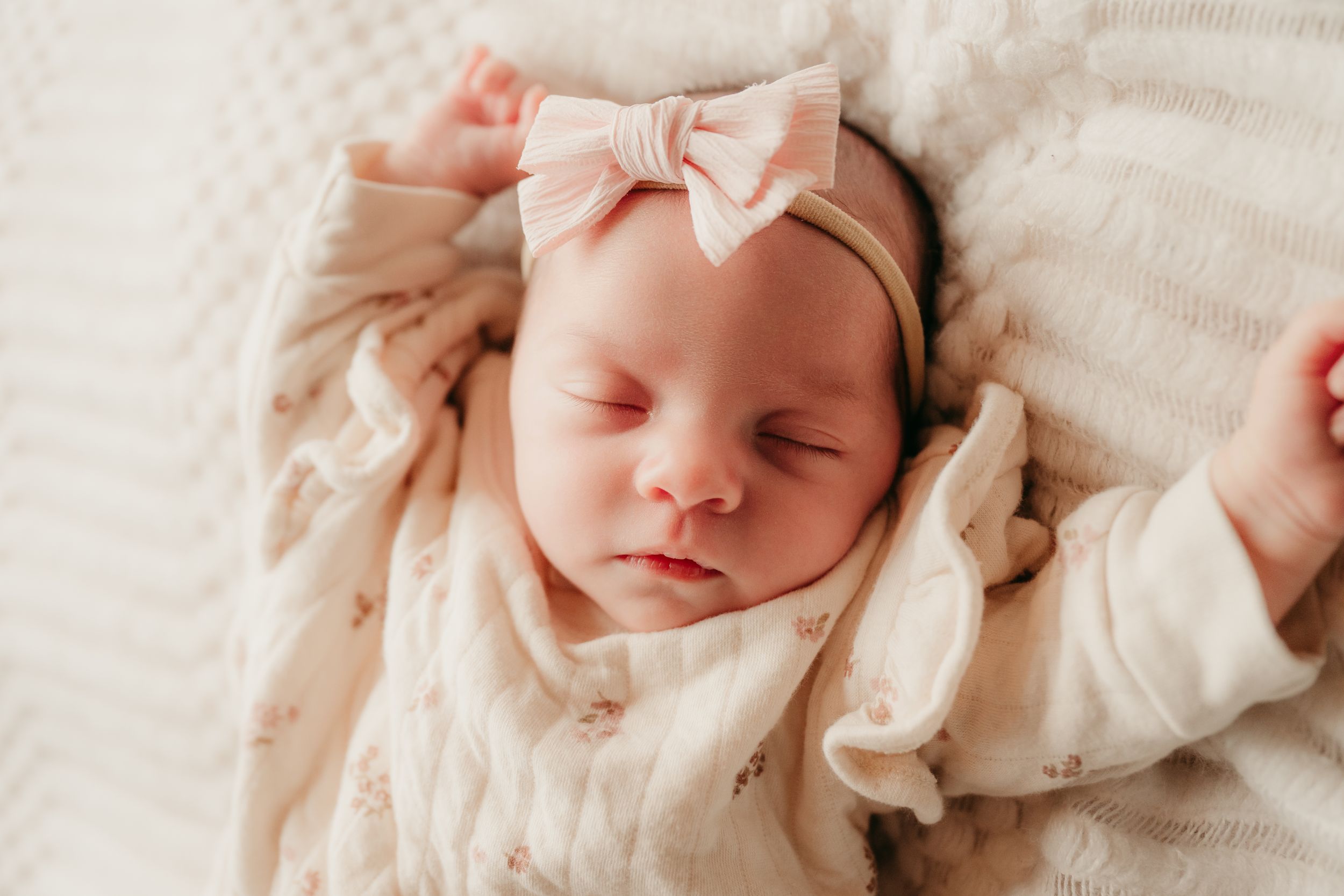 Sleeping baby wearing a pink bow headband rests peacefully on cream colored blanket.
