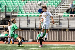 Soccer players in green and white uniforms chase the ball on a turf field with stadium seating visible.