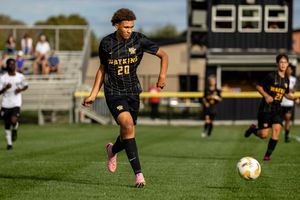 Soccer player in black and gold uniform dribbles ball during competitive match on sunny grass field.