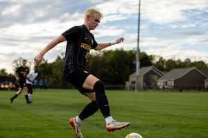 Soccer player in black uniform dribbles ball on grass field during daytime match with houses in background.