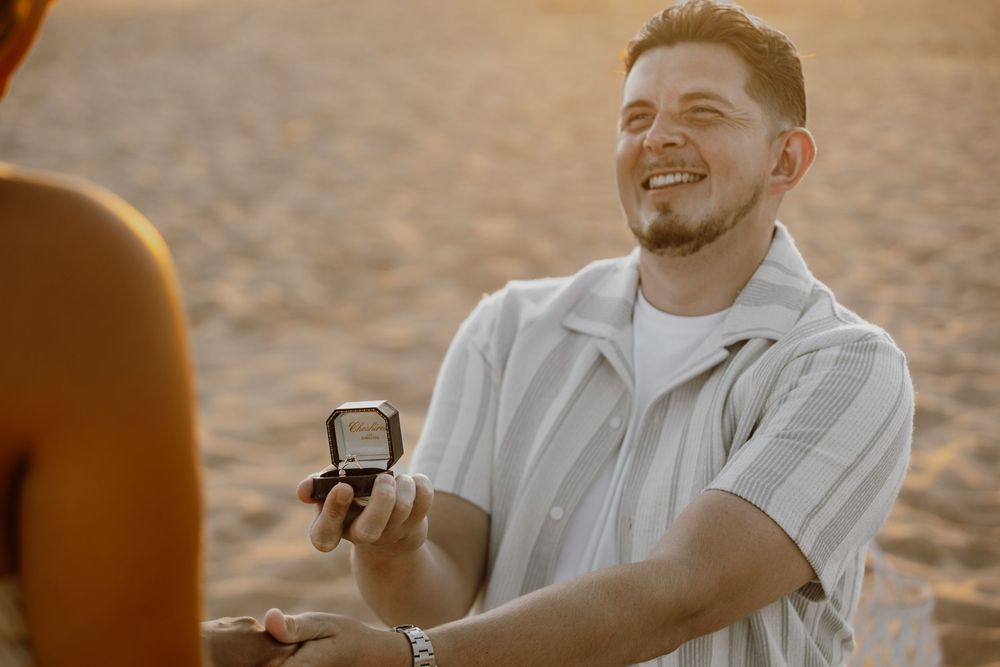 A couple sits together in white outfits during a romantic golden hour beach proposal.