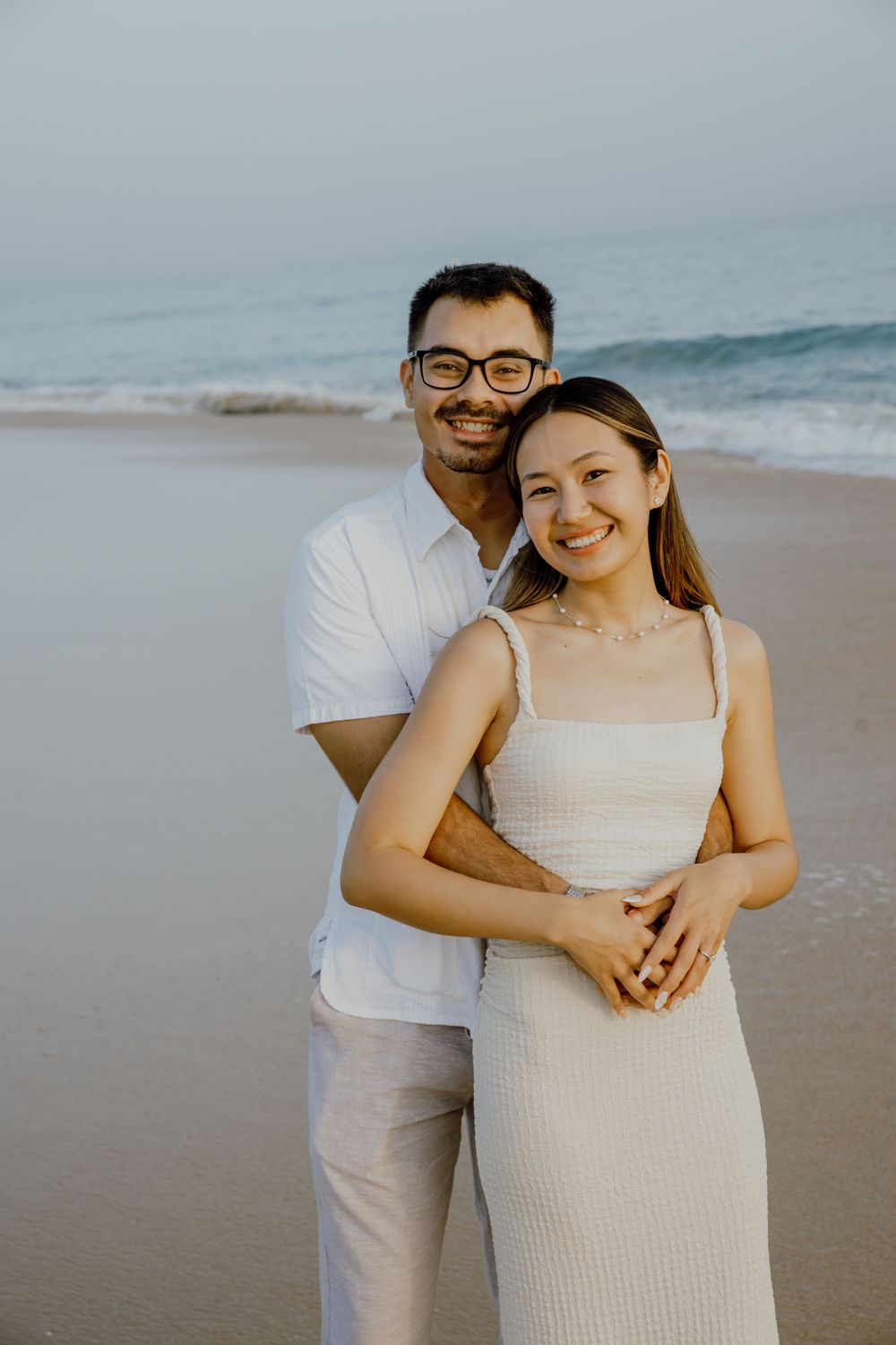 A couple shares a tender moment together on a misty beach while wearing coordinated white outfits.