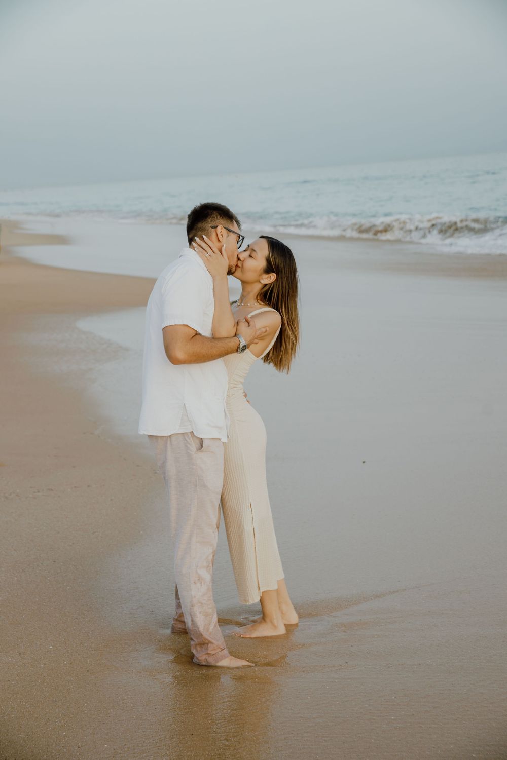 A couple shares a tender moment on a misty beach with soft neutral tones and gentle waves in the background.
