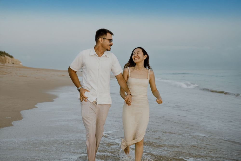 A couple walks hand in hand along a sunny beach shoreline wearing light colored casual outfits.