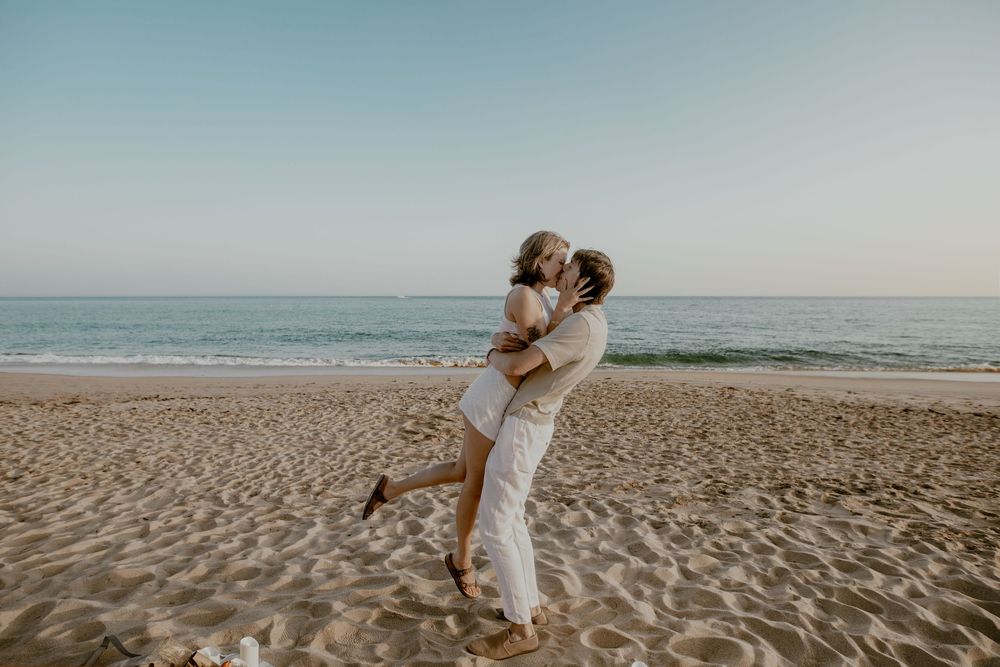 Romantic couple in white outfits embracing on a sandy beach at sunset with ocean waves in the background.