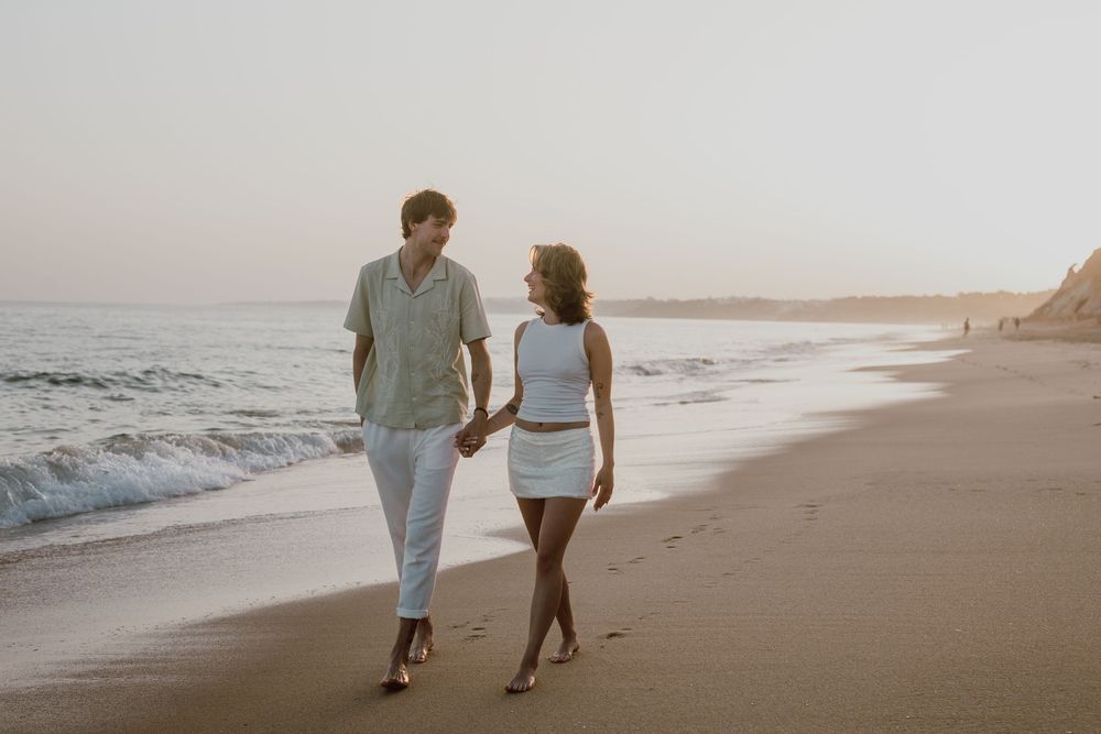 A couple walks hand in hand along a scenic beach at sunset while waves crash on the shore.