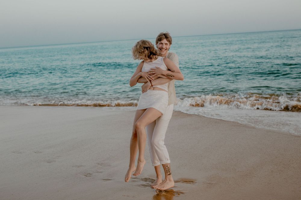 Couple in light colored outfits embracing on a sandy beach with turquoise ocean waters in the background.
