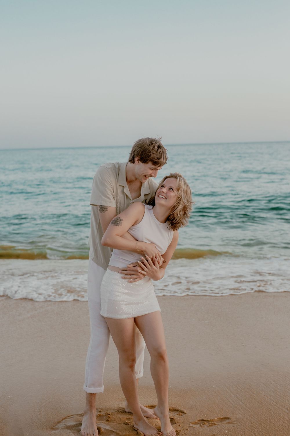 A romantic couple embraces and dances together on a sandy beach with ocean waves in the background at sunset.