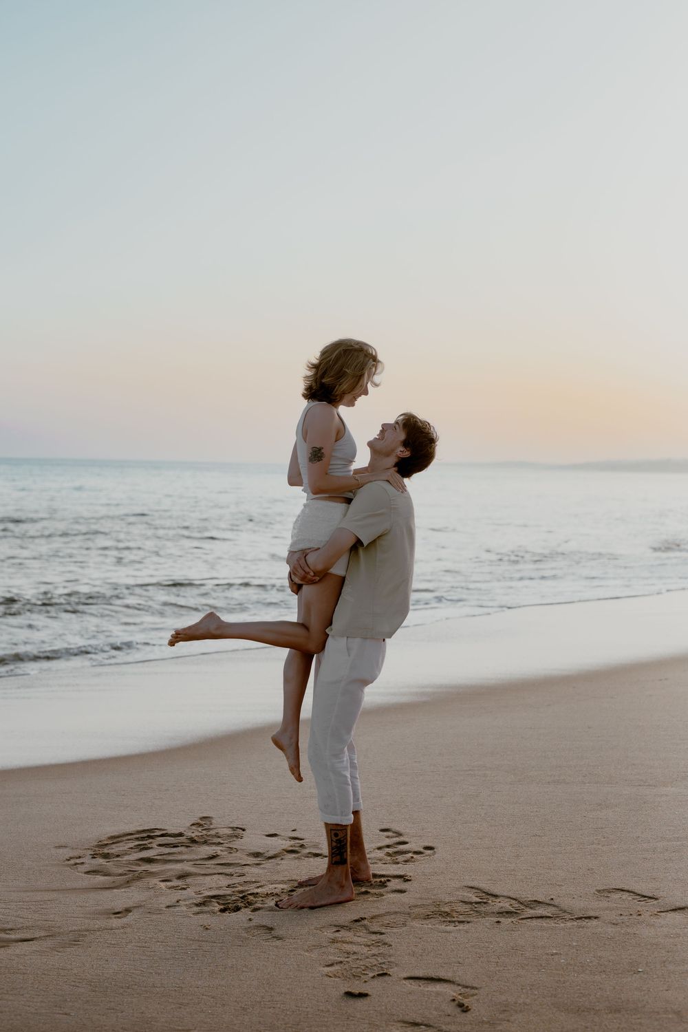 A romantic couple embraces on a sandy beach at sunset in matching white outfits near the ocean waves.