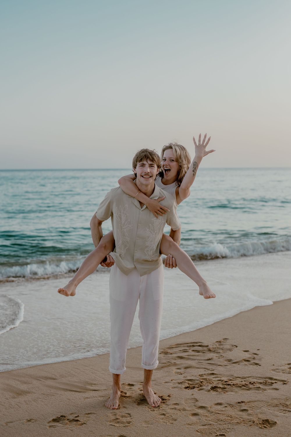 Two people in white outfits playfully pose and dance together on a sandy beach at sunset with ocean waves in background.