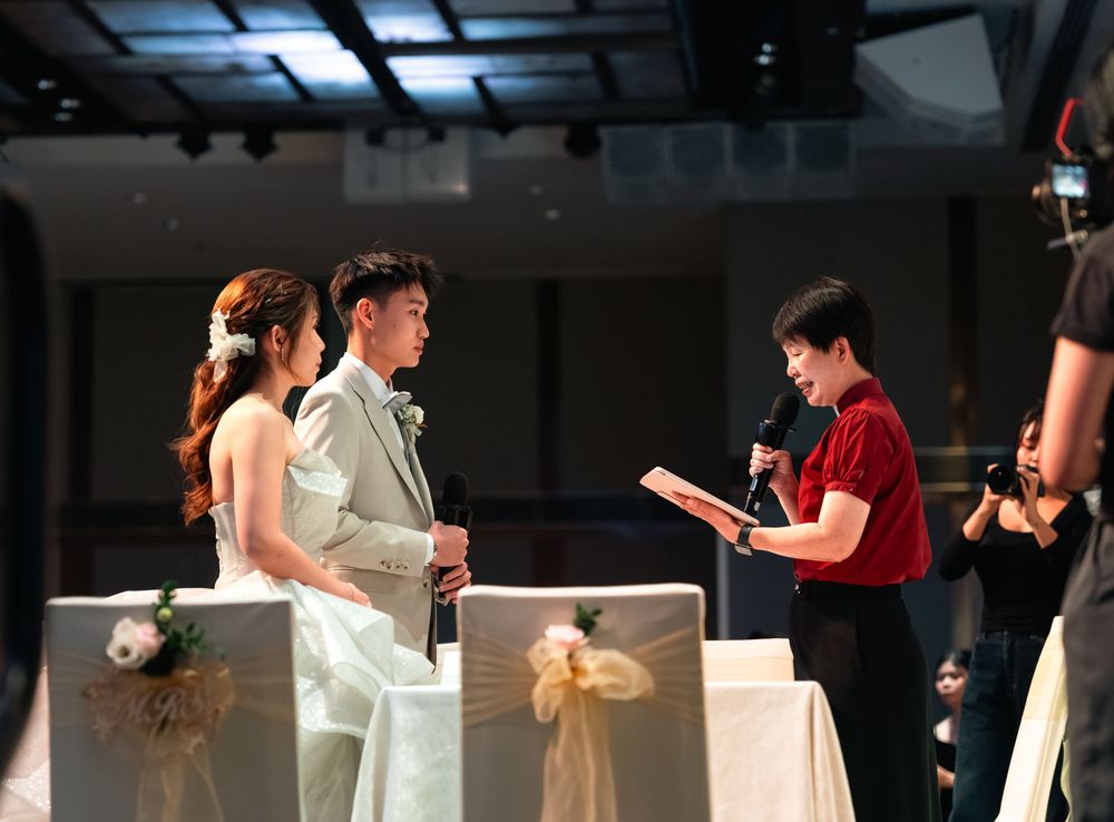 A couple in formal wedding attire stands at an altar while an officiant reads from a book during a ceremony.