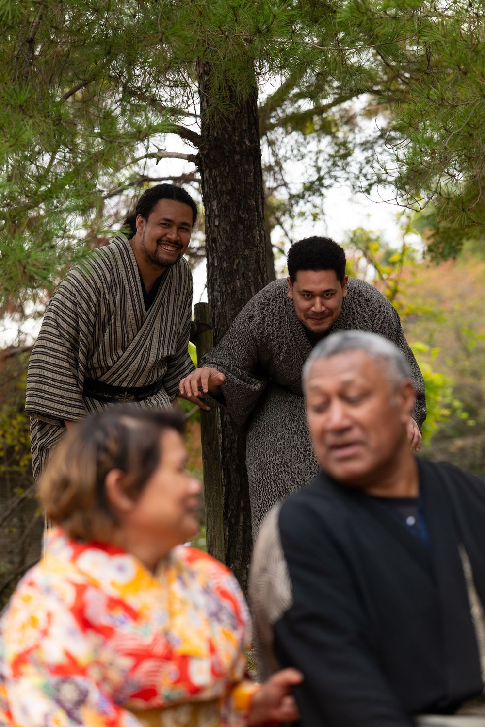Two men in kimono smiling by a pine tree while couple stands in foreground in Japan.