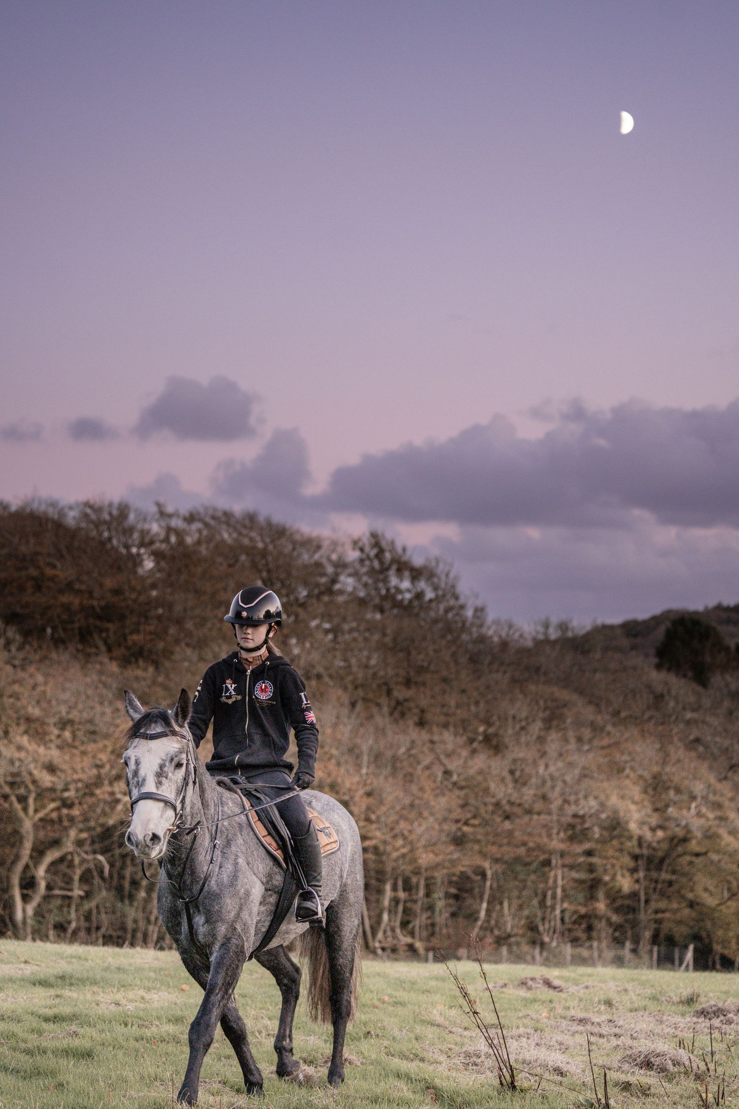 A horse rider on a dappled grey horse in a rural field at dusk with a crescent moon visible in the purple sky.
